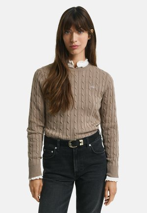 Beige cable-knit sweater with ribbed cuffs, layered over a white ruffled collar shirt, paired with black jeans and a black belt.