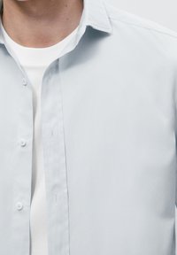 Camicia button-up azzurro chiaro con una texture morbida, vestibilità slim e colletto classico; indossata sopra una t-shirt bianca a girocollo.