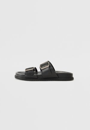 Sort slidesandal med to brede remme med sølvfarvede rektangulære spænder på en flad sål mod en ensartet lys grå baggrund.