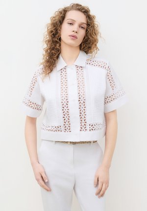 Blonde vrouw met krullend haar die een witte blouse met knopen en bloemmotieven draagt, gecombineerd met een witte broek tegen een effen achtergrond.