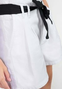 Pantalones cortos blancos de algodón, con un cinturón de tela negra, diseño plisado, bolsillos laterales y un dobladillo limpio.