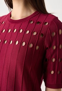 Burgundy neulepaita, jossa on vaaka ribbaus ja tasaisin välein olevat pyöreät leikkaukset. Lyhyet hihat ribbaamalla varustetuilla hihoilla.