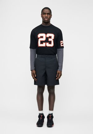 Junger Mann steht, trägt schwarze Shorts, ein schwarzes T-Shirt mit der Nummer 23, darunter ein graues Langarmshirt, weiße Socken und schwarze Turnschuhe.