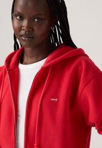 Haut de survêtement zippé rouge en tissu doux avec une poche kangourou, arborant un petit patch logo Levi's sur la poitrine gauche et des cordons de serrage sur la capuche.