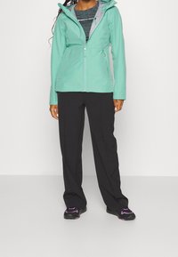 Mintgrön vattentät jacka med dragkedja framtill, justerbar huva och svarta friluftsbyxor. Svarta skor med lila detaljer.