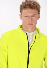 Neon sárga, zipzáras kabát magas gallérral, sima textúrával és fekete cipzár részletekkel. Fehér ing felett viselve.