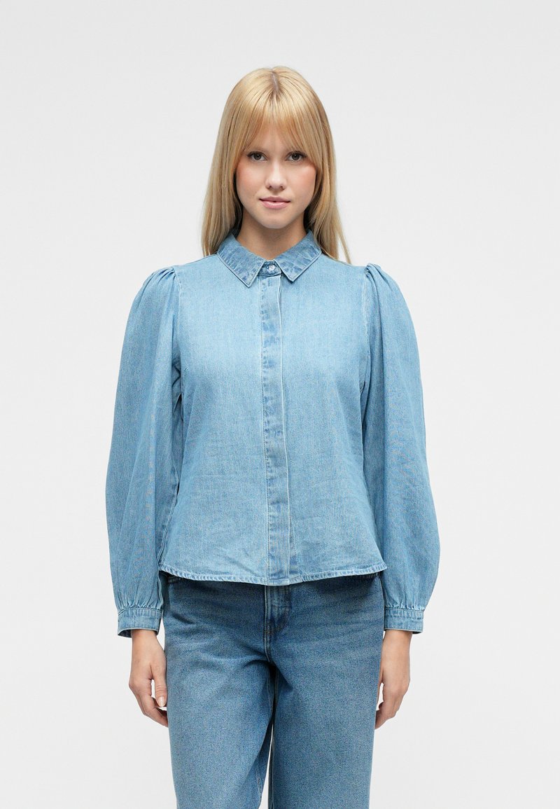 Jasnoniebieska koszula dżinsowa z bufkami na rękawach, spiczastym kołnierzem i zapięciem na guziki z przodu. Gładka tekstura, luźny krój.