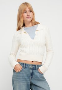 Pulover crem cu torsade, cu gât în V și design cu guler; are manșete striate și o lungime scurtă, purtat împreună cu blugi albastru deschis.