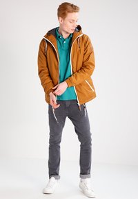 Giacca leggera marrone con cappuccio, cerniere bianche e polsini elasticizzati. Abbinata a un polo teal, jeans grigi e sneakers bianche.