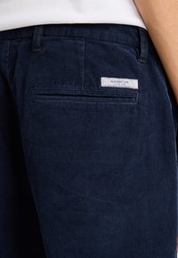 Pantalones de pana azul marino con un bolsillo trasero que presenta una etiqueta de tela, "ELEMENT CO." Tela texturizada, de superficie acanalada y diseño estructurado.