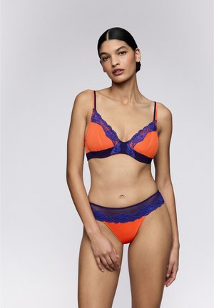 Vrouw met donker haar draagt een oranje bh en bijpassend onderbroekje met donkerblauwe kanten accenten, staand tegen een effen lichtgrijze achtergrond.
