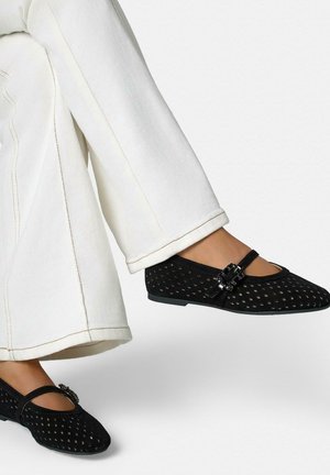 Zapatos planos negros tipo Mary Jane perforados con hebilla decorativa, usados con pantalones blancos de pierna ancha en pies elevados.