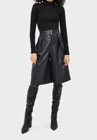 Kobieta ubrana w czarną bluzkę z długim rękawem zapinaną na perłowe guziki, czarne skórzane spódnice kulotowe o wysokim stanie oraz czarne muszkieterki na obcasie.