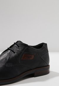 Chaussure en cuir noir à texture lisse, avec un patch logo marron, un design élégant et des lacets foncés. Accentuée par des coutures contrastantes.