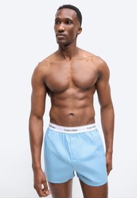 Mężczyzna o muskularnej budowie, bez koszulki, ubrany w jasnoniebieskie bokserki Calvin Klein, stojący i patrzący na bok na jednolitym tle.