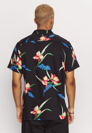 Homme aux cheveux courts et bouclés portant une chemise noire à manches courtes avec des motifs floraux rouges et roses ainsi que des feuilles bleues, vu de dos.