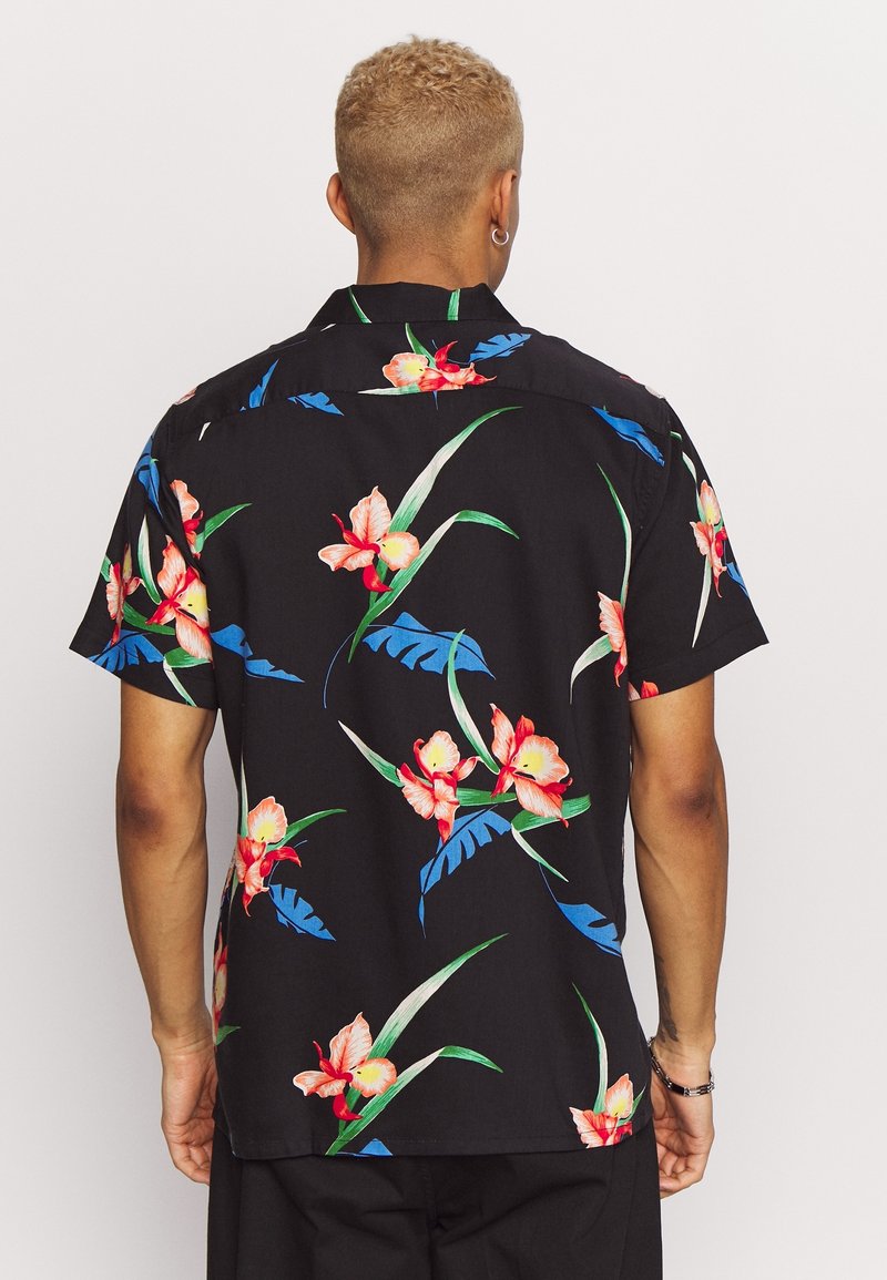 Homme aux cheveux courts et bouclés portant une chemise noire à manches courtes avec des motifs floraux rouges et roses ainsi que des feuilles bleues, vu de dos.