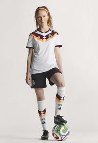 Maillot de soccer blanc avec des accents rouges, jaunes et noirs, associé à un short noir et des chaussettes montantes à motifs géométriques. Chaussures noires.