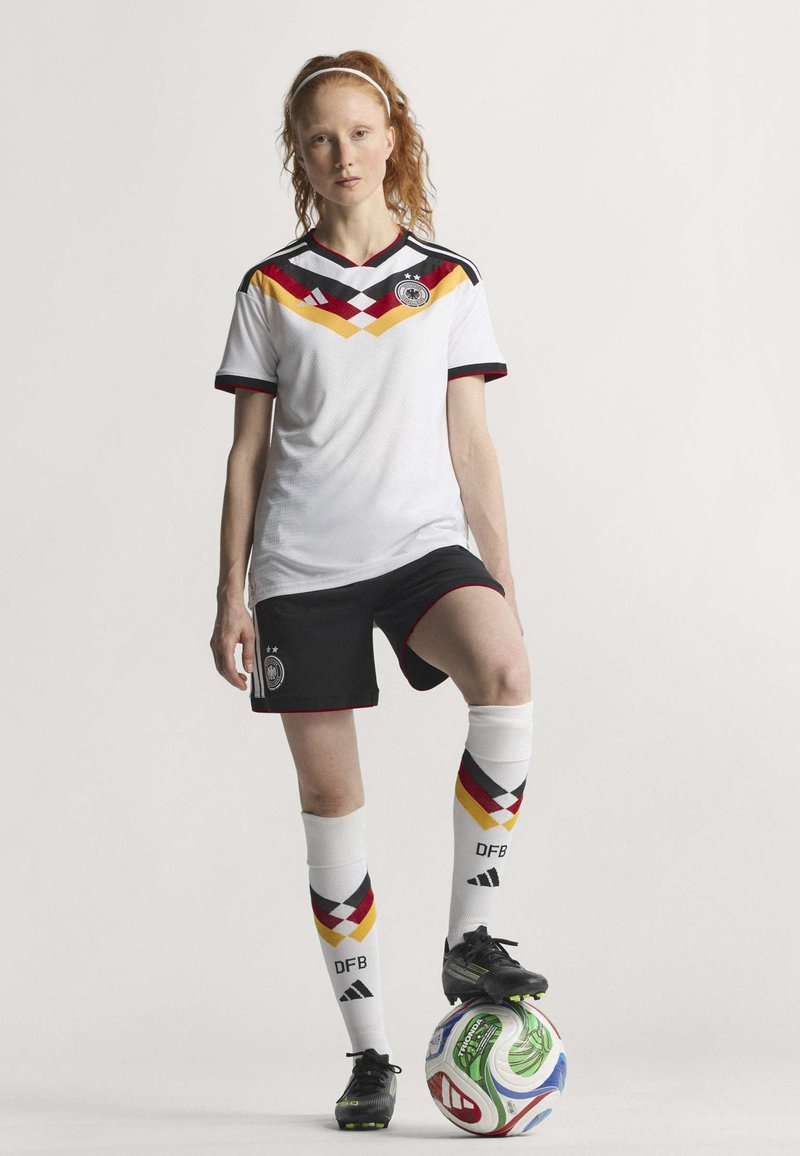 Maillot de soccer blanc avec des accents rouges, jaunes et noirs, associé à un short noir et des chaussettes montantes à motifs géométriques. Chaussures noires.