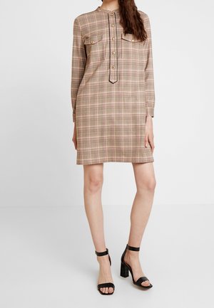 Woman wearing a beige plaid long-sleeve dress with gold buttons and black high-heeled sandals, standing against a plain background.