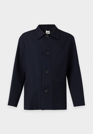 Homme portant une veste noire ample, un pantalon noir et des chaussures noires tenant une grande pochette bleu marine sur fond blanc.