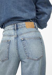 Gros plan d'une personne portant un jean en denim bleu clair taille haute avec des poches arrière visibles et un patch de marque en cuir marron sur la ceinture.