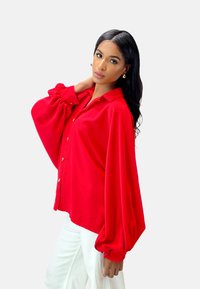 Red long-sleeve blouse with a relaxed fit, button front, and voluminous sleeves. Smooth fabric with a slight sheen. Worn with white pants.