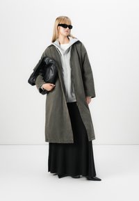 Lungo cappotto in denim verde oliva con bottoni, felpa grigia sotto, gonna nera e accessori neri tra cui una borsa e occhiali da sole.