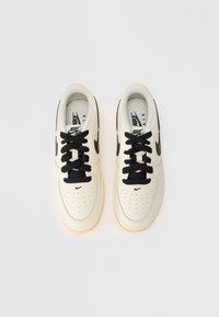 Sapatilhas Nike Air em couro creme com detalhes pretos, apresentando atacadores redondos pretos, caixa de biqueira perfurada e sola de borracha translúcida.