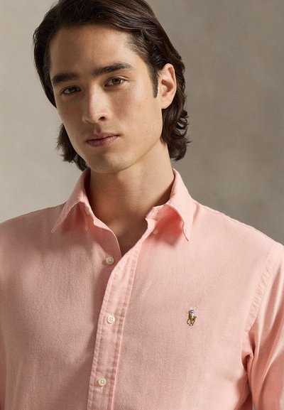 Jauns vīrietis ar vidēja garuma tumšiem matiem valkā gaiši rozā pogājamu kreklu ar mazu izšūtu polo spēlētāja logo uz krūtīm.