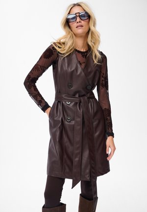 Blonde Frau mit großen Sonnenbrillen, transparenter schwarzer Bluse mit Blumenmuster und langen Ärmeln, ärmellosem braunen ledernen Kleid mit Gürtel und dunklen Hosen mit Stiefeln.