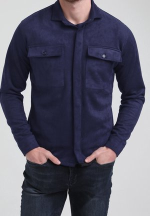 Man in een navyblauw shirt met lange mouwen, voorzien van klepzakken op de borst en donkerblauwe spijkerbroek, handen in de voorzakken, staand tegen een effen achtergrond.