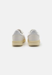 Weiße Ledersneaker mit hellgelben Sohlen. Merkmale sind eine abgerundete Form, textile Akzente und das "New Balance"-Logo an der Ferse.