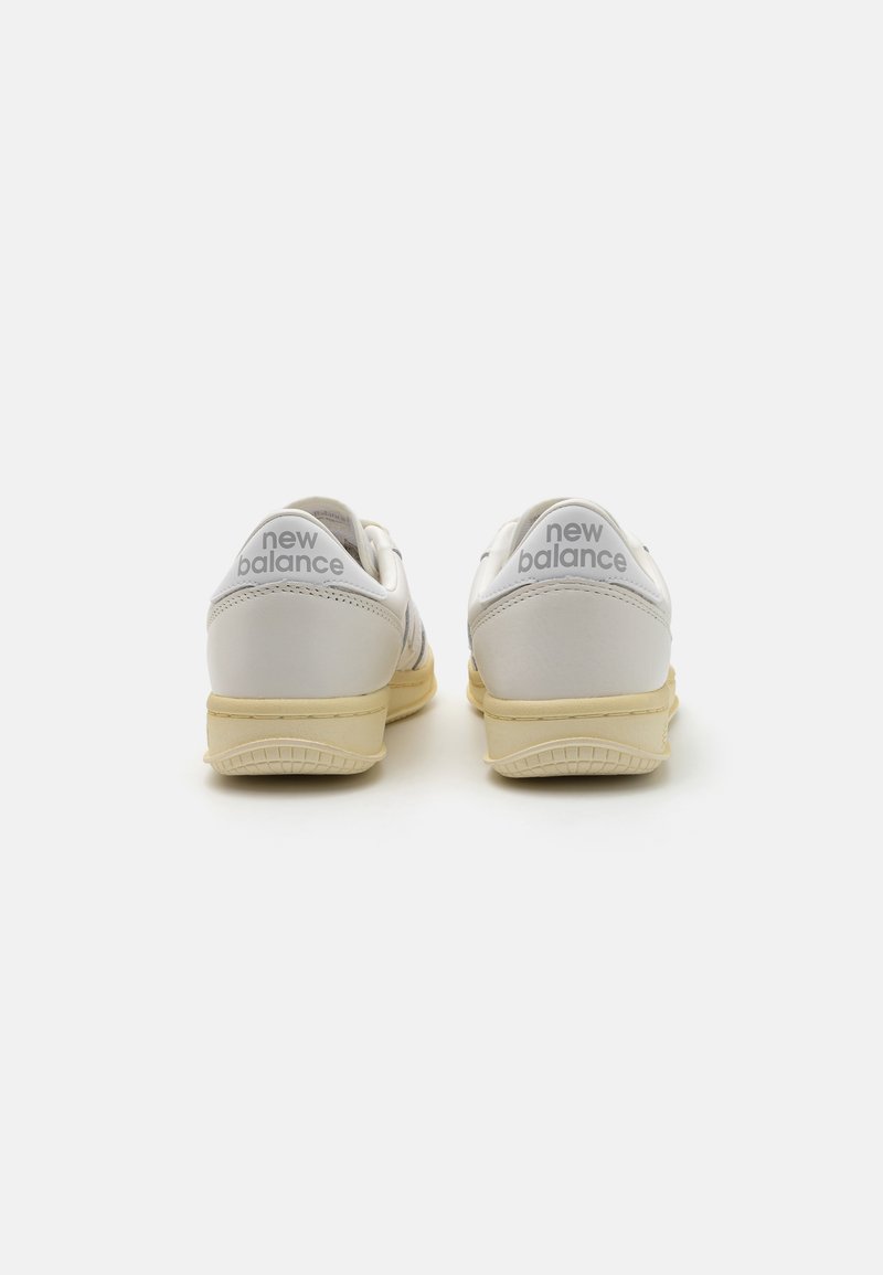 Weiße Ledersneaker mit hellgelben Sohlen. Merkmale sind eine abgerundete Form, textile Akzente und das "New Balance"-Logo an der Ferse.