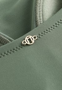 Nahaufnahme eines grünen Stoffes mit glatter Textur, Netzdetail und einem goldfarbenen runden Logo-Anhänger mit Nähten entlang des Randes.
