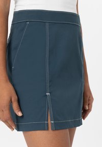 Modrá džínsová sukně s přiléhavým střihem, kontrastním bílým prošíváním a postranním rozparkem pro větší detaily. Hladká textura s viditelným pasem.