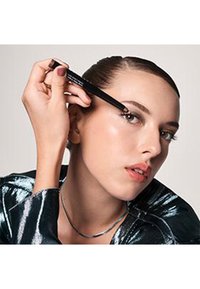 Matita eyeliner nera tenuta da una persona, mentre si applica sulla palpebra superiore. L'individuo indossa un top metallizzato lucido e una delicata collana.