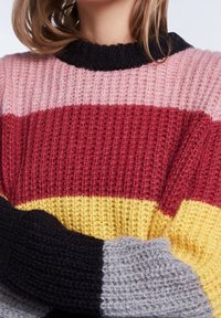 Donna che indossa un maglione lavorato a maglia con strisce orizzontali di rosa, rosso, giallo, nero e grigio, con le braccia incrociate.