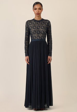 Frau trägt ein langärmeliges schwarzes Spitzenoberteil mit rundem Ausschnitt und einen schwarzen, plissierten bodenlangen Rock, steht vor einem schlichten Hintergrund.