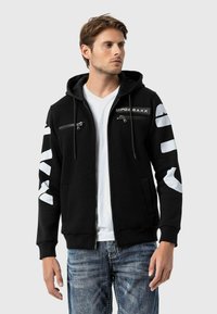 Schwarzer Hoodie mit Reißverschluss-Taschen vorne, Kordelzug-Kapuze und weißen grafischen Akzenten an den Ärmeln und auf der Brust. Hergestellt aus weichem Stoff.