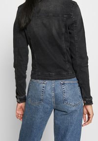 Svart denimjacka med långa ärmar, figurnära design, synlig sömnad och knappade manschetter. Bärs med ljusblå jeans med hög midja.