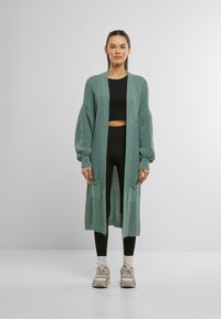 Long cardigan tricoté en teal clair avec une ouverture à l'avant, des poignets côtelés et deux poches avant. Porté par-dessus un crop top noir et des leggings.