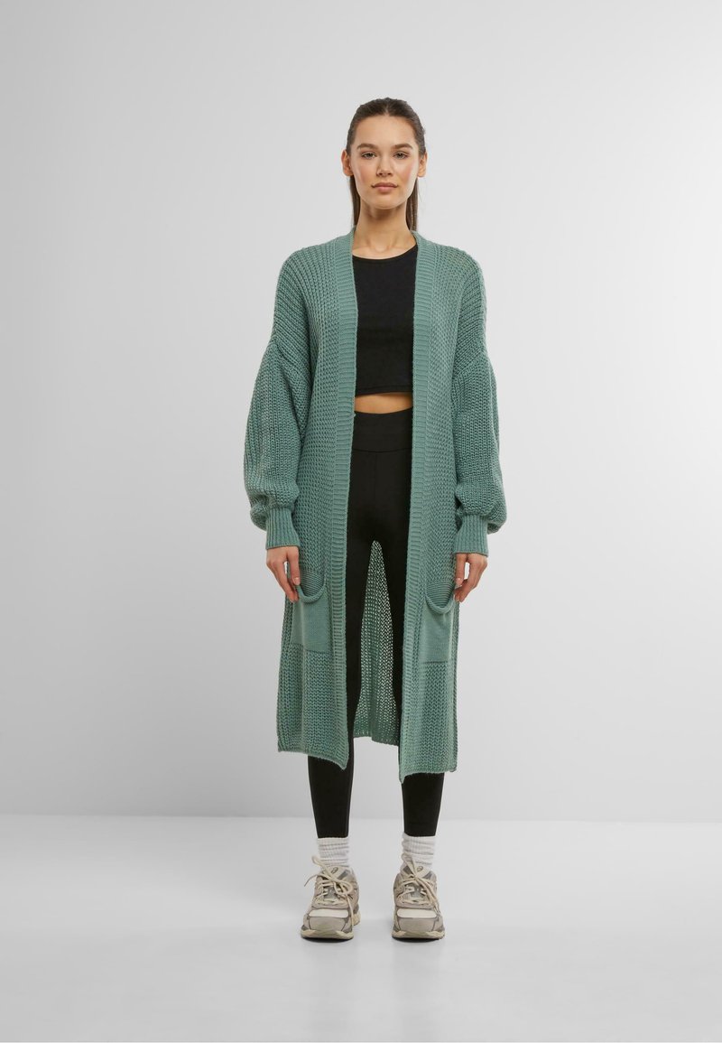 Long cardigan tricoté en teal clair avec une ouverture à l'avant, des poignets côtelés et deux poches avant. Porté par-dessus un crop top noir et des leggings.
