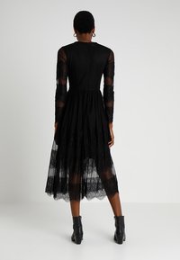 Robe noire avec de longues manches en dentelle transparente, un corsage ajusté et une jupe en dentelle à couches. Dispose d'une fermeture éclair au dos et d'une coupe longueur cheville.