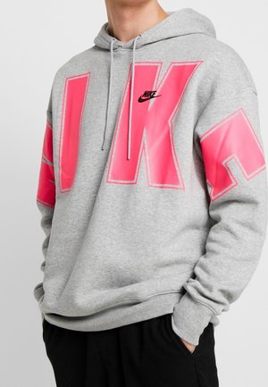 Person iført en grå Nike-hættetrøje med store lyserøde bogstaver "NIKE" tværs over brystet og ærmerne, hænderne i lommerne.