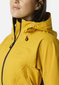 Giacca impermeabile gialla con collo alto, zip frontale, cappuccio regolabile e logo ricamato. Presenta dettagli neri e zip sicure.