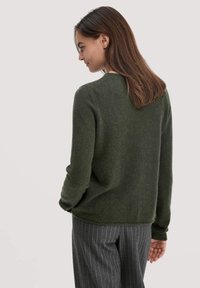Dunkelgrüner Strickpullover mit rundem Halsausschnitt und langen Ärmeln, mit einer strukturierten Oberflächenbeschaffenheit. Kombiniert mit gestreiften Hosen, von hinten betrachtet.