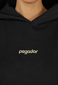 Juodas džiusas, pagamintas iš minkštos medžiagos, su priekyje esančia kišene ir raišteliu ant kepurės. Išsiuvinėtas "pegador" logotipas kontrastinga smėlio spalva.