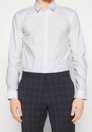 Homme portant une chemise blanche cintrée à boutons et un pantalon marine foncé à carreaux, debout avec les bras relâchés sur les côtés.