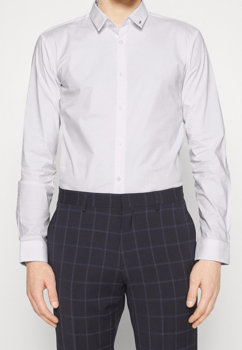 Homme portant une chemise blanche cintrée à boutons et un pantalon marine foncé à carreaux, debout avec les bras relâchés sur les côtés.